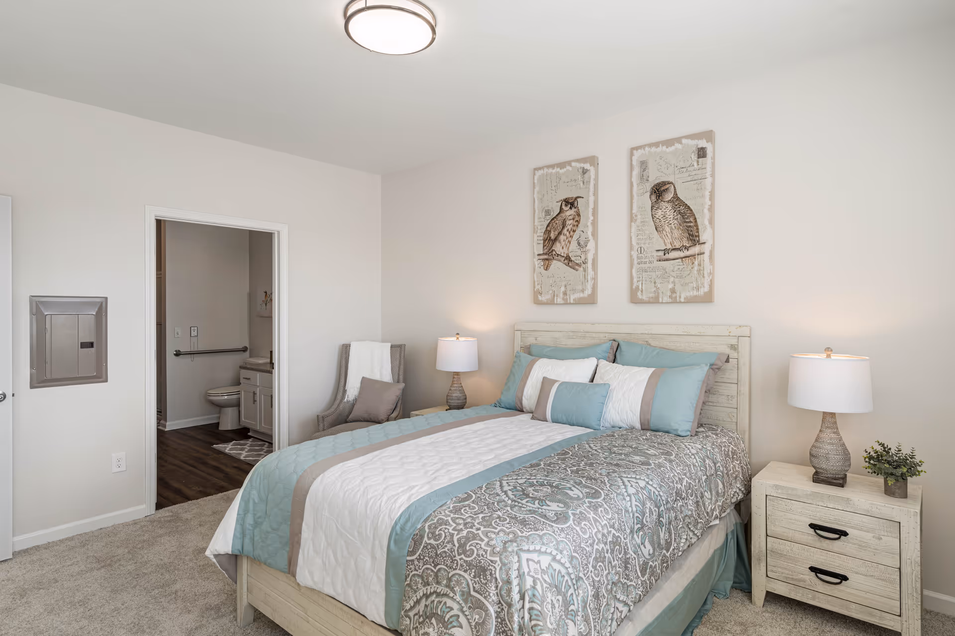 A bedroom with a neatly made bed featuring a patterned comforter in shades of teal, white, and beige. Two framed owl artworks hang above the headboard. There is a nightstand with a lamp and a small plant on the right side of the bed. A gray armchair with a pillow and a white throw blanket is positioned near the open doorway leading to a bathroom with a visible toilet and vanity. The room has light-colored walls and carpeted flooring.