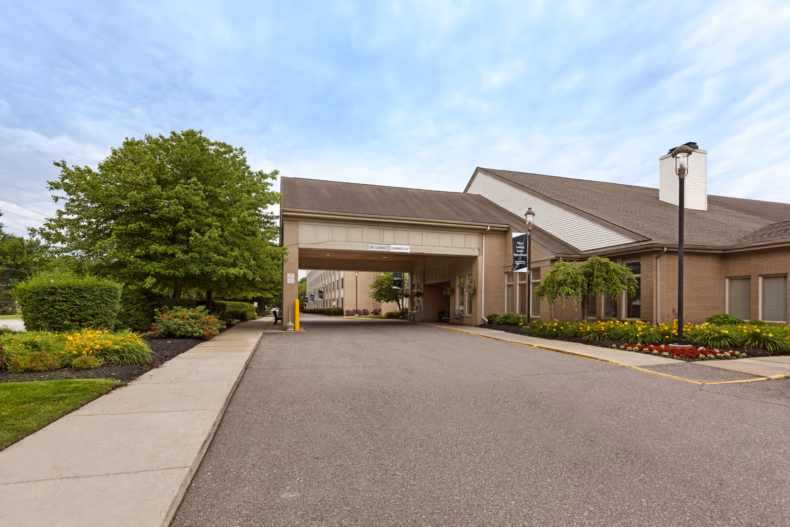 Exterior view of Independence Village of Plymouth showing the driveway entrance with a covered drop-off area, surrounded by well-maintained landscaping including trees, bushes, and flower beds under a partly cloudy sky.