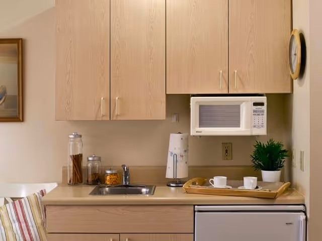 A small kitchen area with light wood cabinets, a countertop with a sink, a microwave mounted above the counter, a small refrigerator below, and a tray with two white cups and a teapot. There are also jars with snacks, a paper towel holder, a green potted plant, and a wall clock.