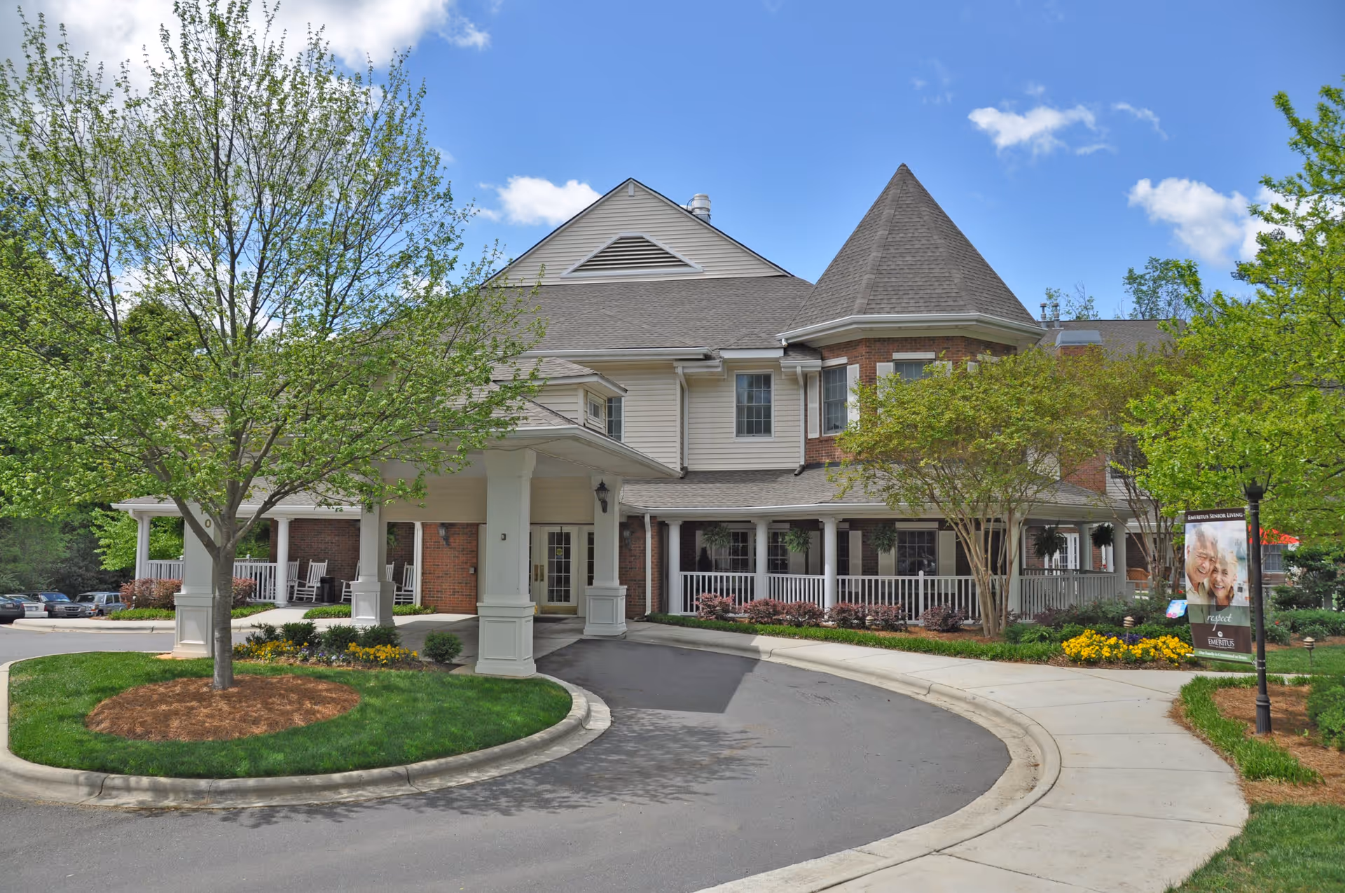 Exterior view of Charter Senior Living of Charlotte, showing a large building with a covered entrance, surrounded by green trees and landscaped gardens under a blue sky with some clouds.