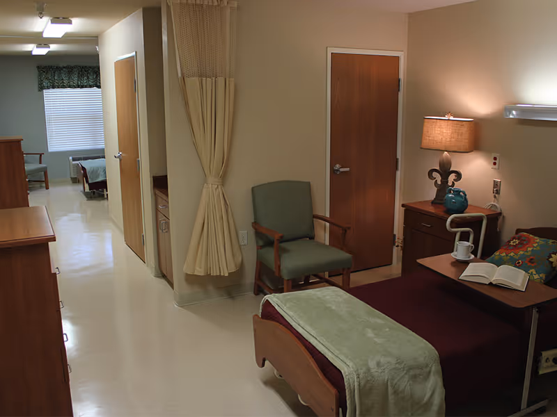 Furnished senior living bedroom with a made bed, overbed table and lamp, a chair, privacy curtain, and a hallway to another room.