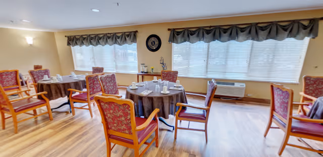 A dining room with multiple round tables covered with brown tablecloths, each set with white cups and saucers. The room has wooden chairs with red patterned cushions, large windows with blinds and valances, and a wooden floor. There is a wall clock and an air conditioning unit below the windows.