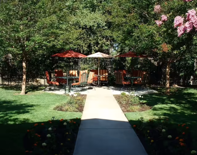 Outdoor patio area with several tables and chairs under red and beige umbrellas, surrounded by green trees and flowering plants, with a concrete walkway leading to the seating area.