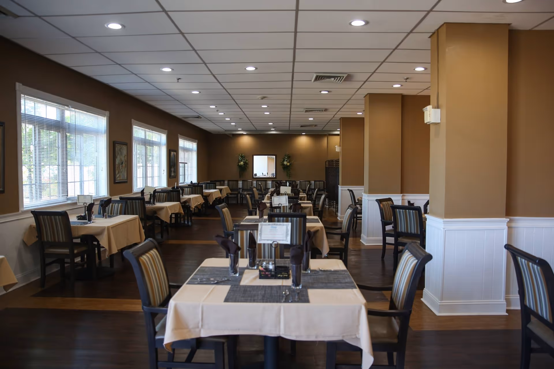A spacious dining room with multiple tables covered in beige tablecloths and set with dark napkins and utensils. The room has large windows on one side allowing natural light to enter, brown walls with white wainscoting, and recessed ceiling lights. Chairs with striped upholstery are arranged around the tables.
