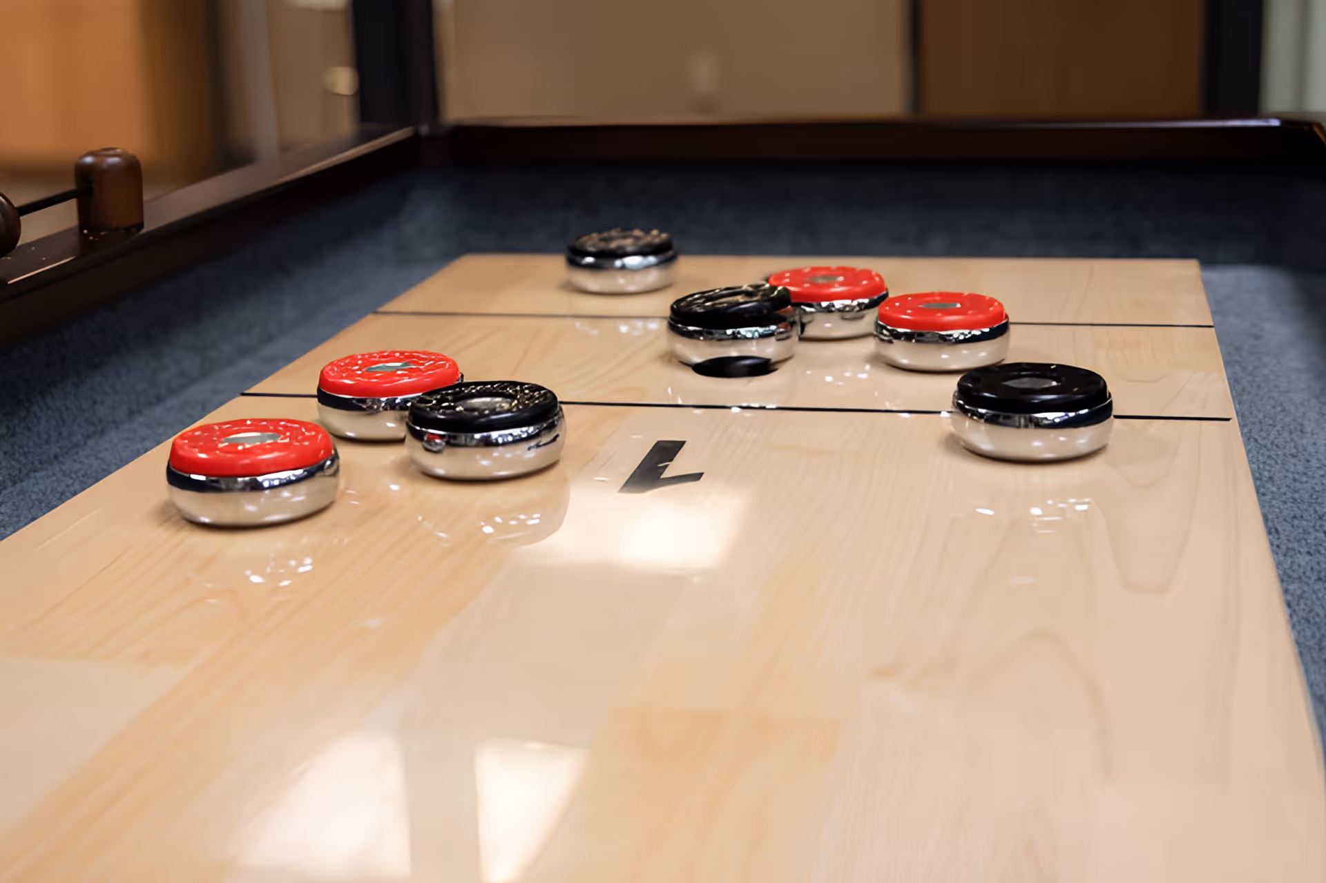 Close-up view of a shuffleboard table with red and black pucks arranged on the wooden playing surface inside a room.