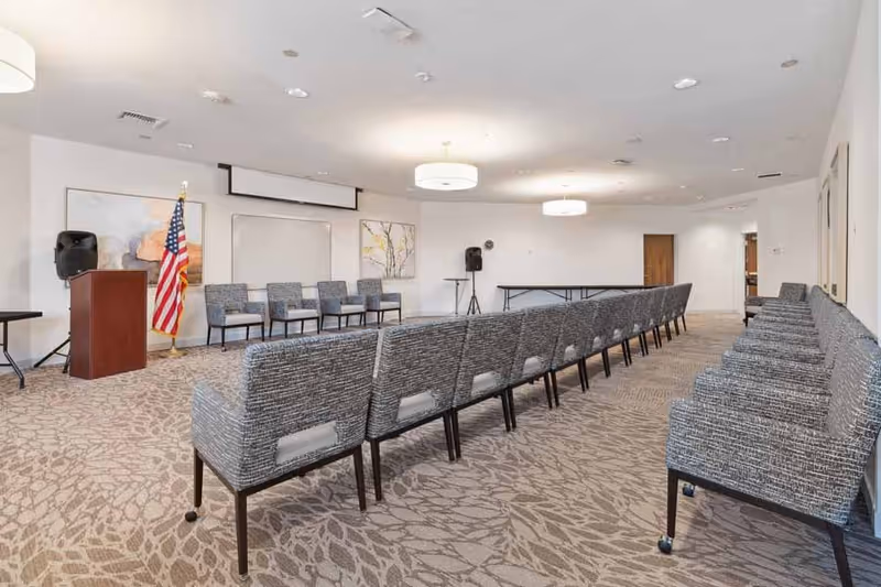 A conference or meeting room with rows of gray upholstered chairs facing a podium with an American flag beside it. The room has beige patterned carpet, white walls, ceiling lights, and speakers on stands.