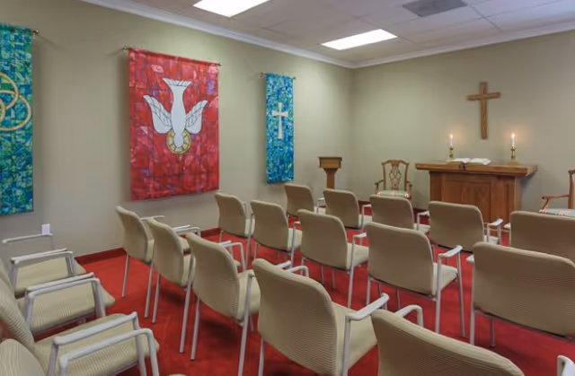 Interior view of a small chapel or worship room with rows of beige chairs facing a wooden altar. The altar has an open book and two lit candles. A wooden cross is mounted on the wall behind the altar. Colorful banners with religious symbols hang on the walls. The room has a red carpet and beige walls with overhead fluorescent lighting.