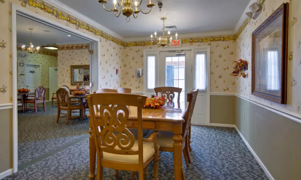 A dining room in a senior living facility with a wooden table and chairs. The room has patterned wallpaper with a fruit motif, a carpeted floor, and two chandeliers hanging from the ceiling. There is a door with windows letting in natural light and a framed picture on the wall. Another dining area is visible through an open doorway.