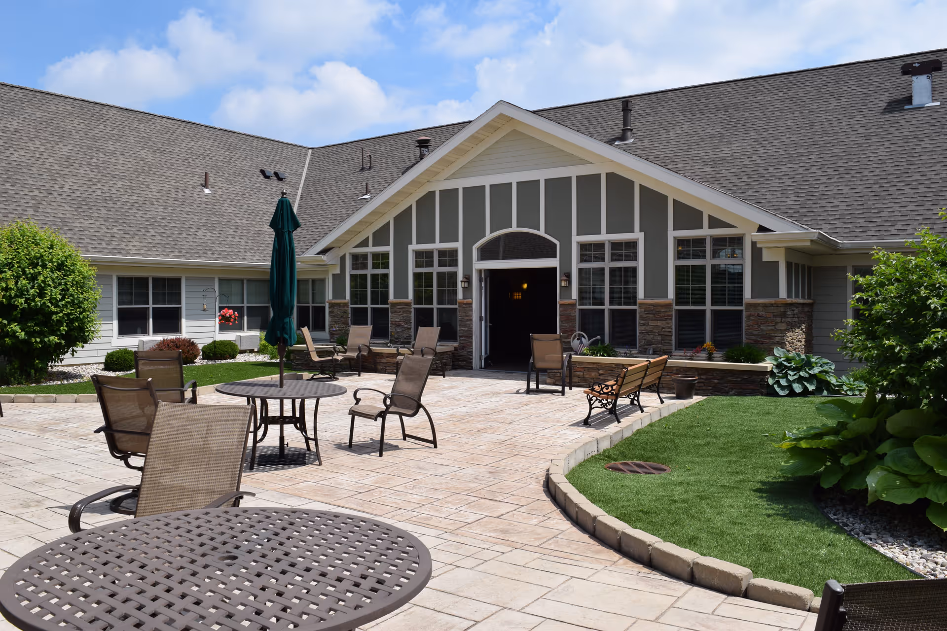 Outdoor patio with tables, chairs, benches and an umbrella in front of a single-story senior living building entrance.