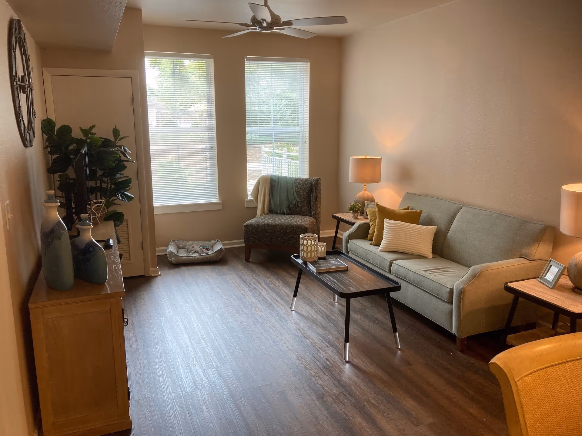 A cozy living room with a light green sofa adorned with pillows, a patterned armchair, a wooden coffee table with decorative items, two side tables with lamps, a ceiling fan, and a TV on a wooden stand. Two large windows with blinds allow natural light to fill the room, and a pet bed is placed near the windows.