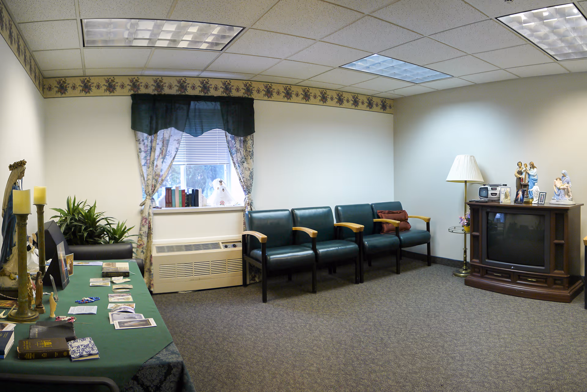 A small sitting room with four green chairs lined up against a wall, a window with floral curtains, a table covered with a green cloth displaying books and religious items, a wooden TV stand with a TV and religious figurines, and a floor lamp in the corner.