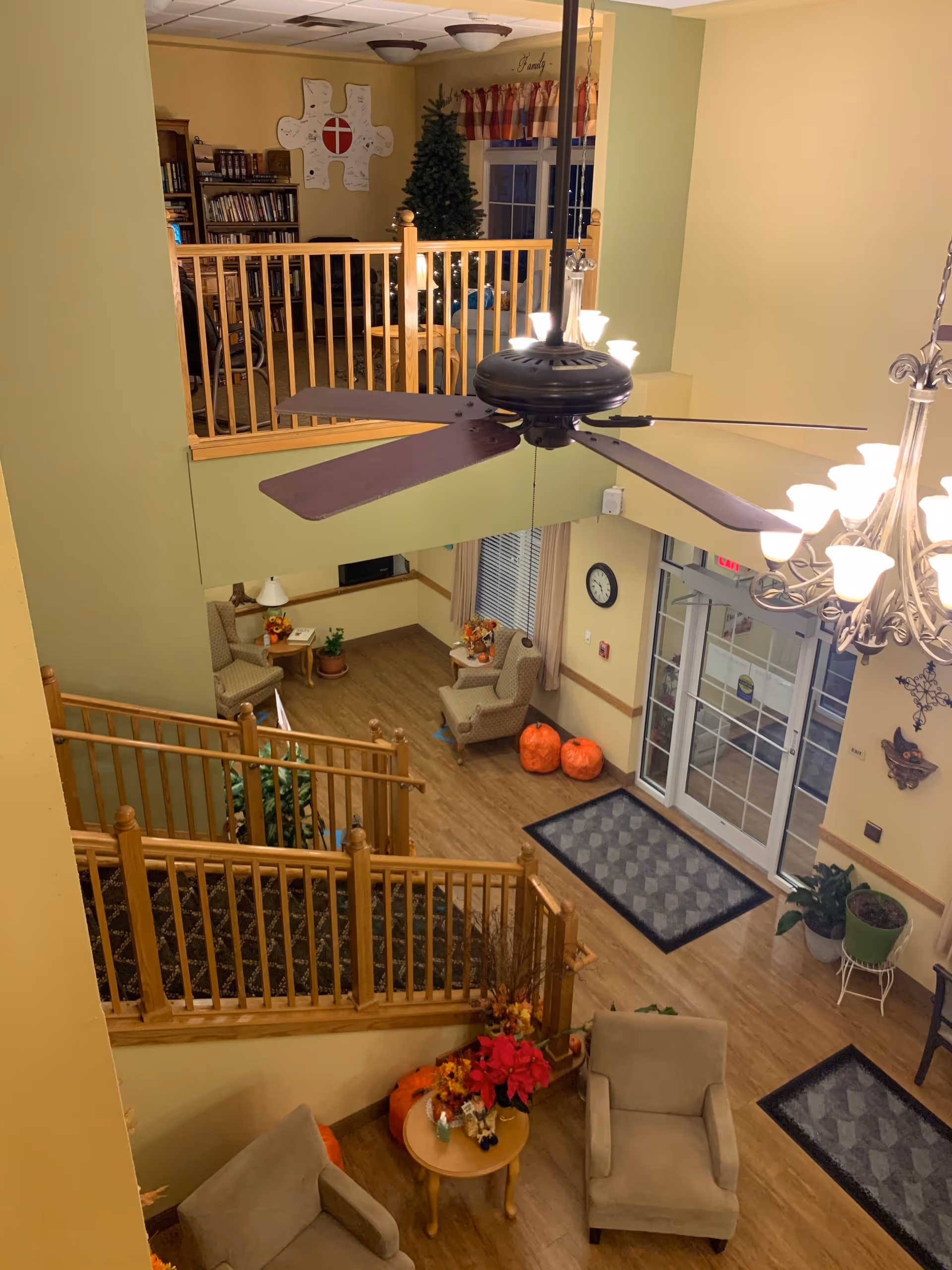 Interior view of a senior living facility lobby area with a high ceiling, ceiling fan, and chandelier lighting. The space includes comfortable armchairs, small tables with floral decorations, pumpkins, and potted plants. A staircase with wooden railings leads to an upper level with bookshelves and a Christmas tree. The entrance door has glass panels and there are wall clocks and decorative items on the walls.