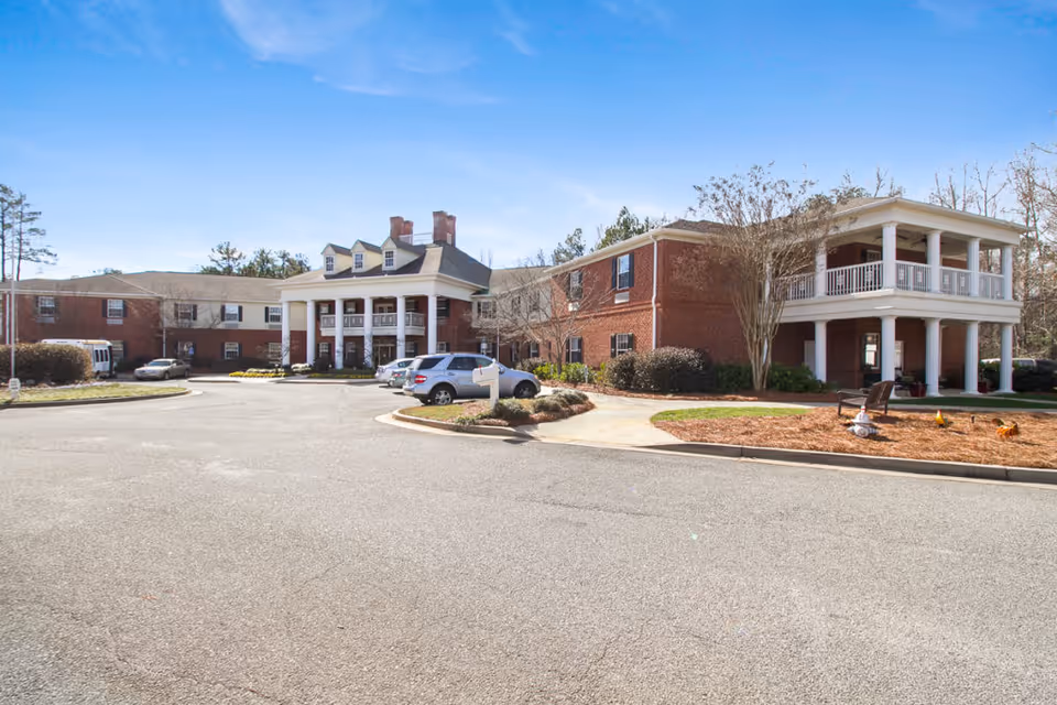 Exterior view of a two-story brick senior living facility with white columns and balconies, a parking area with several cars, and a clear blue sky.