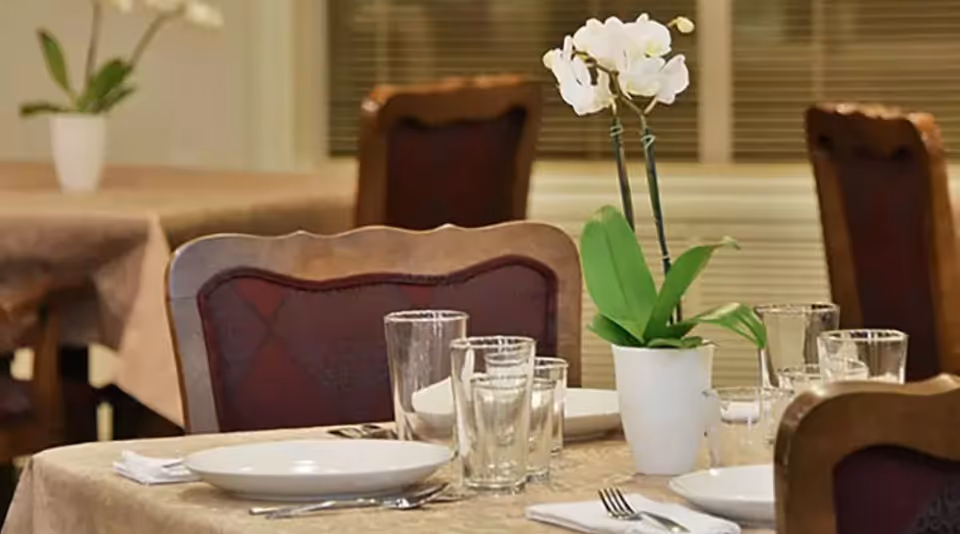 A dining table set with white plates, clear drinking glasses, silverware, and a white potted orchid plant as a centerpiece. The table is covered with a beige tablecloth and surrounded by wooden chairs with red cushioned backs. Additional tables and chairs are visible in the background.