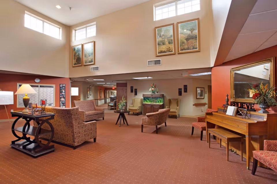 A spacious senior living facility common area with high ceilings and large windows near the ceiling. The room is furnished with patterned armchairs, a wooden piano with a mirror above it, a table with a lamp and decorative items, and an aquarium against the far wall. The walls are decorated with framed paintings and there is a carpeted floor in a warm reddish-brown tone.