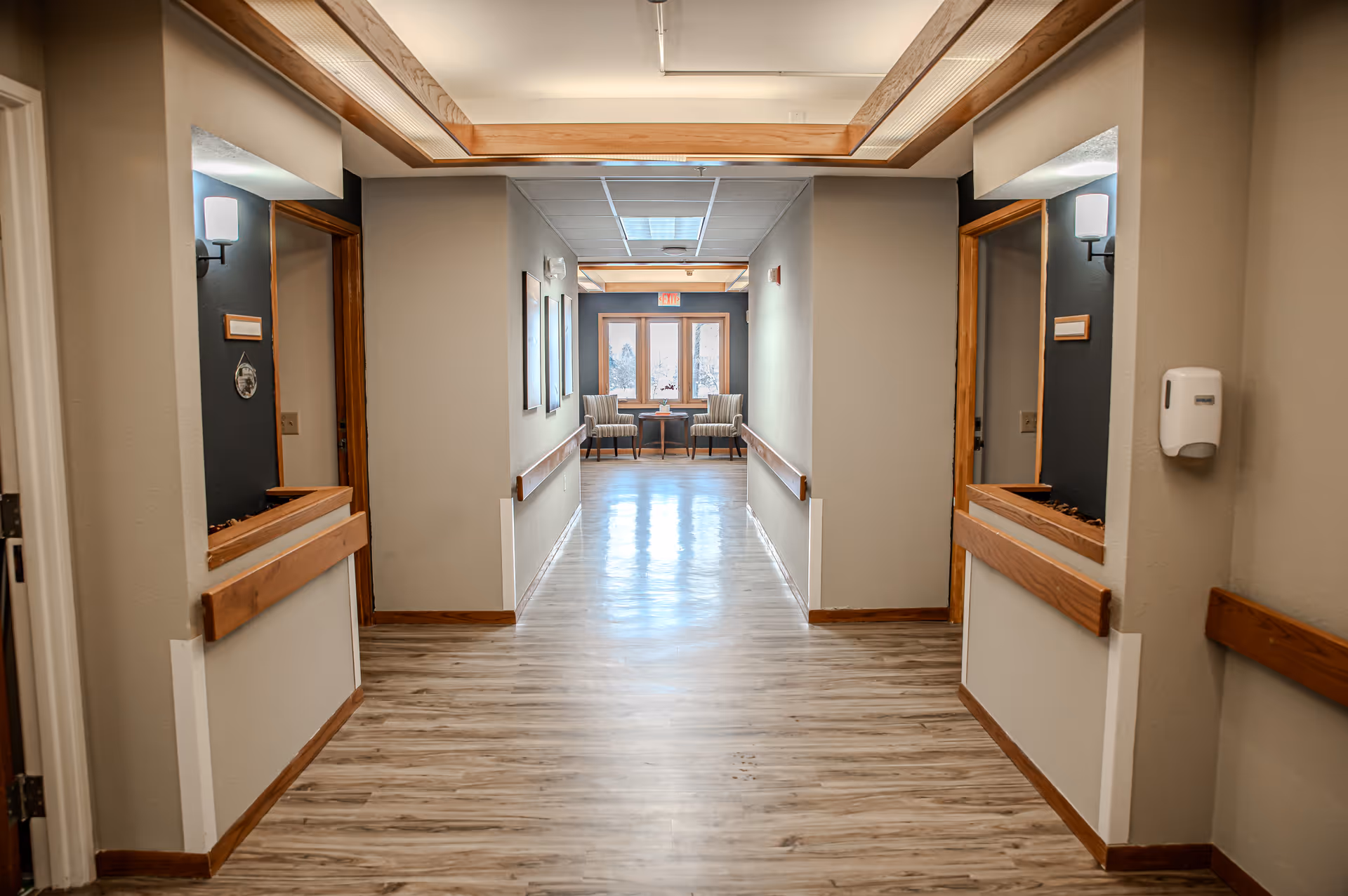 A clean, well-lit hallway in a senior living facility with light wood flooring, beige walls, wooden trim, and handrails on both sides. At the end of the hallway, there are two chairs and a small table near a window with natural light coming through. Wall sconces provide additional lighting along the hallway.