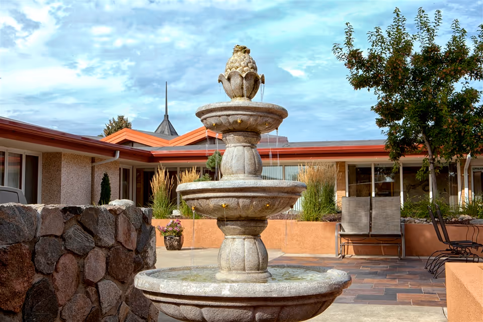 Outdoor courtyard area at Missouri Slope featuring a three-tier stone fountain with water flowing from each tier. Surrounding the fountain are stone walls, potted plants, patio chairs, and a tree, with a building and partly cloudy sky in the background.