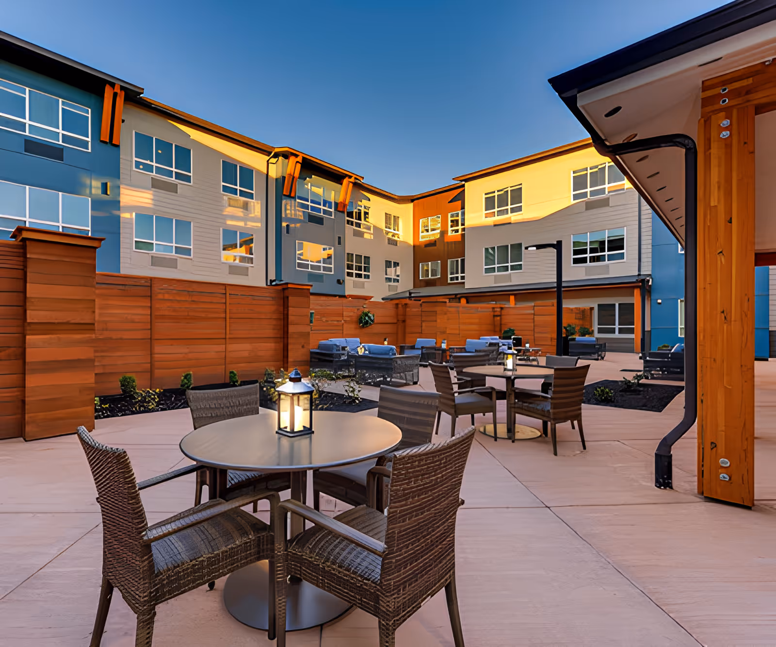Outdoor courtyard patio with round tables, wicker chairs, lanterns and lounge seating surrounded by a three-story residential building.