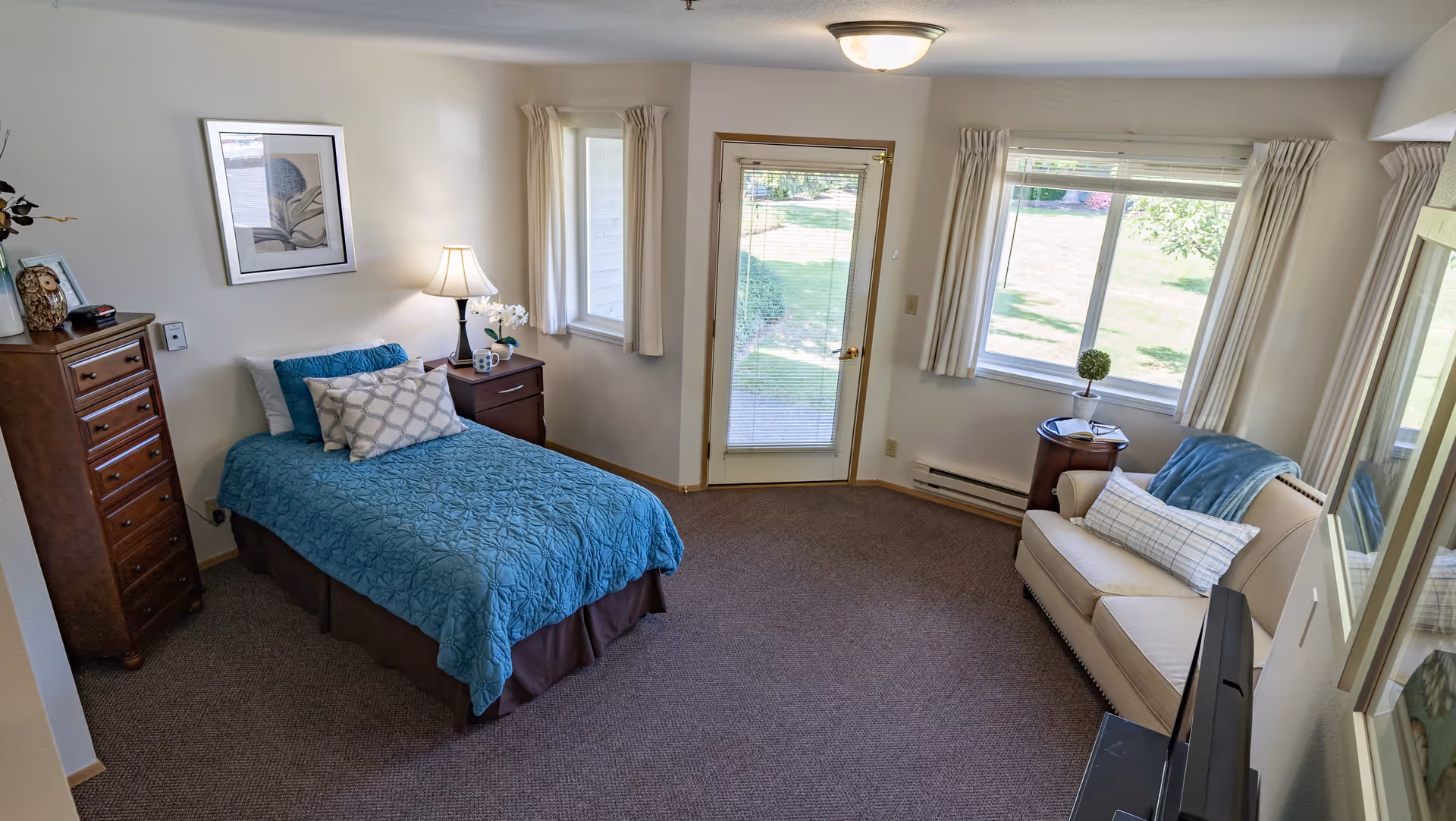 A cozy bedroom in a senior living facility with a single bed covered in a blue quilt and decorative pillows. Next to the bed is a wooden nightstand with a lamp and a small plant. There is a tall wooden dresser against the wall. The room has two windows and a glass door leading outside, allowing natural light to fill the space. A beige loveseat with pillows and a blue throw blanket is positioned near the windows. The room has carpeted flooring and light-colored walls with framed artwork.