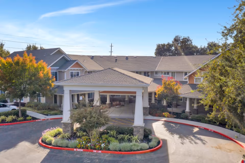 Front exterior view of The Commons At Elk Grove facility showing a covered entrance with a circular driveway, landscaped garden with flowers and shrubs, and a multi-story building with a gray roof and beige siding under a clear blue sky.