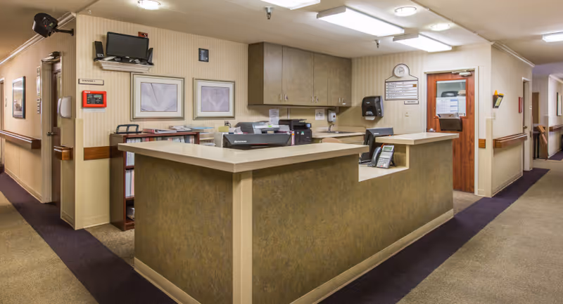 Reception desk area inside Anaheim Terrace Care Center with a counter, computer monitors, office supplies, and cabinets mounted on the wall. The space has beige walls, carpeted floors, framed artwork, and a hallway extending to the right.