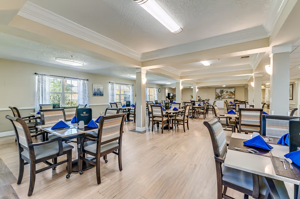 A bright and spacious dining room in a senior living facility with multiple tables set with blue napkins, silverware, and menus. The room has large windows with sheer curtains, light-colored walls, wooden flooring, and ceiling lights. Chairs with cushioned seats and backs surround the tables.