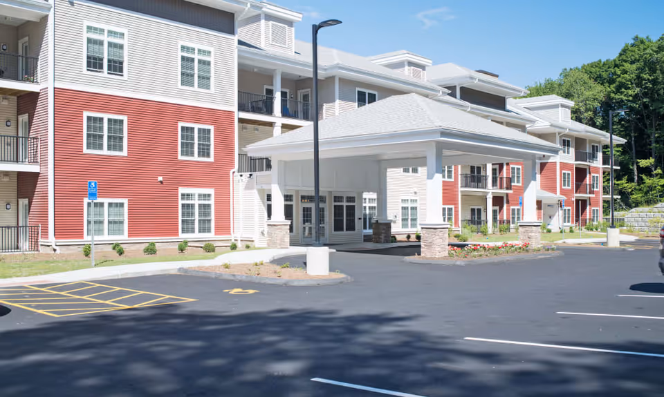 Exterior view of a multi-story senior living facility building with red and beige siding, white trim, and multiple balconies. The entrance features a covered drop-off area with stone pillars. There is a parking lot with marked spaces and a handicap parking sign in front of the building. Trees and greenery are visible in the background under a clear blue sky.