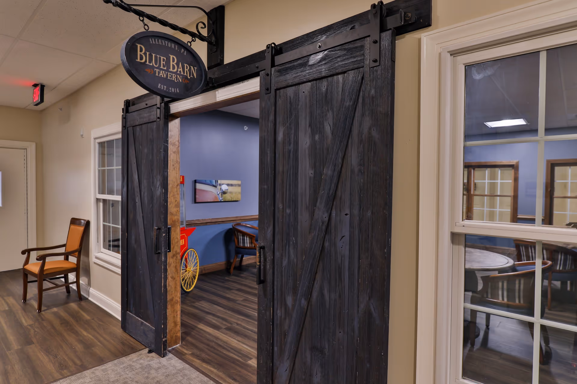 Interior hallway of Heather Glen Senior Living showing a rustic sliding barn door entrance to the Blue Barn Tavern. The hallway has wood flooring, beige walls, a wooden chair with an orange cushion, and a window looking into a room with tables and chairs.
