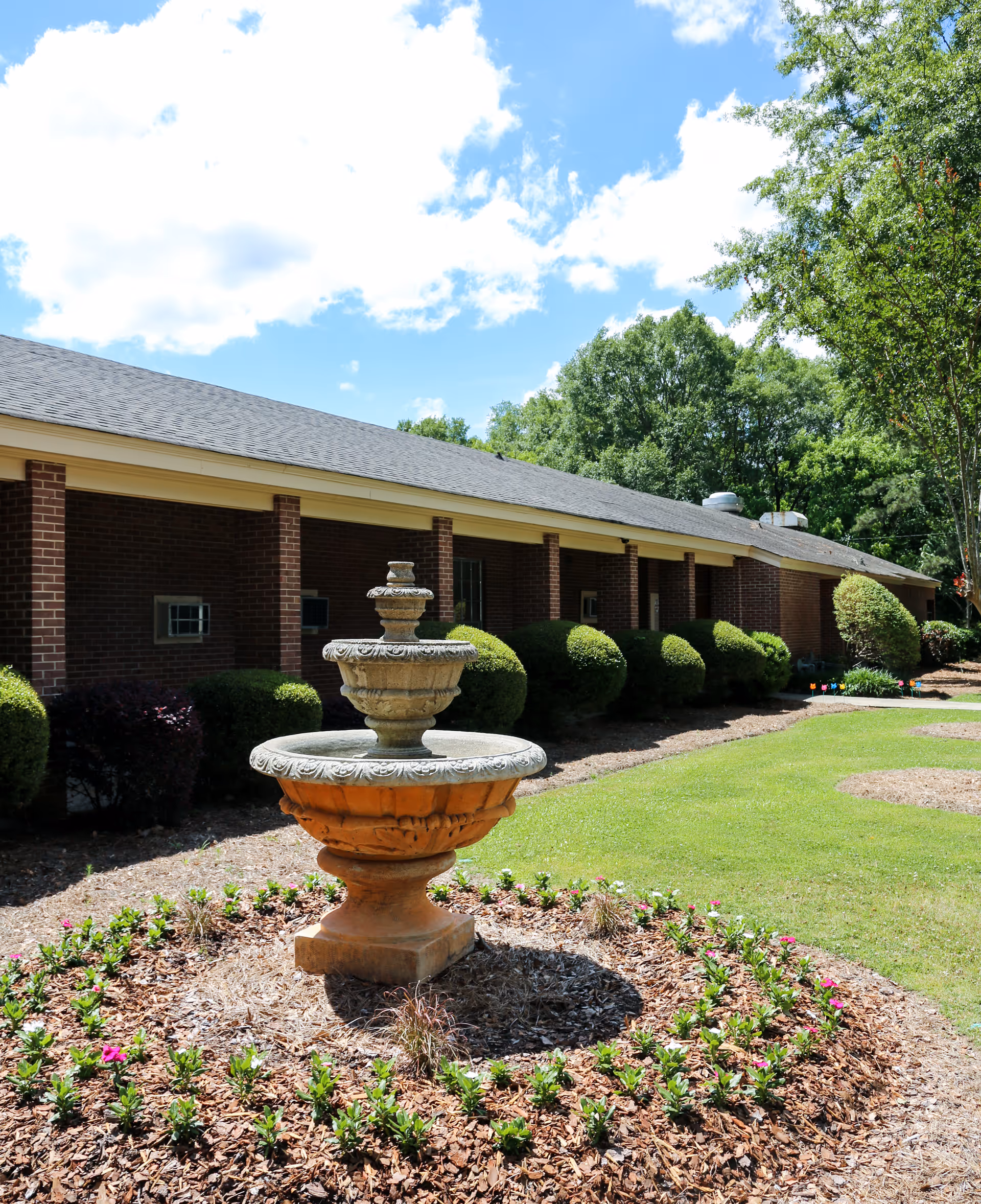 Outdoor view of a senior living facility with a decorative stone fountain in the foreground surrounded by a circular flower bed. The building is a single-story brick structure with a covered walkway and neatly trimmed bushes. The sky is partly cloudy with blue patches visible.