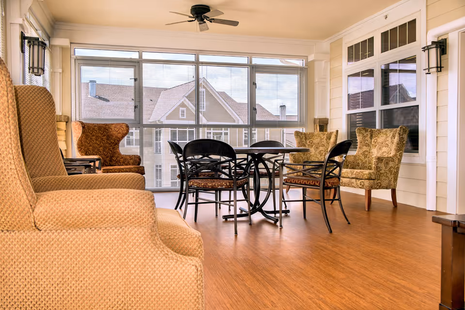 A bright and cozy indoor seating area with large windows showing the exterior of a building. The room features a round table with four black chairs and several upholstered armchairs in beige and patterned fabric. The floor is wooden, and a ceiling fan is mounted above.