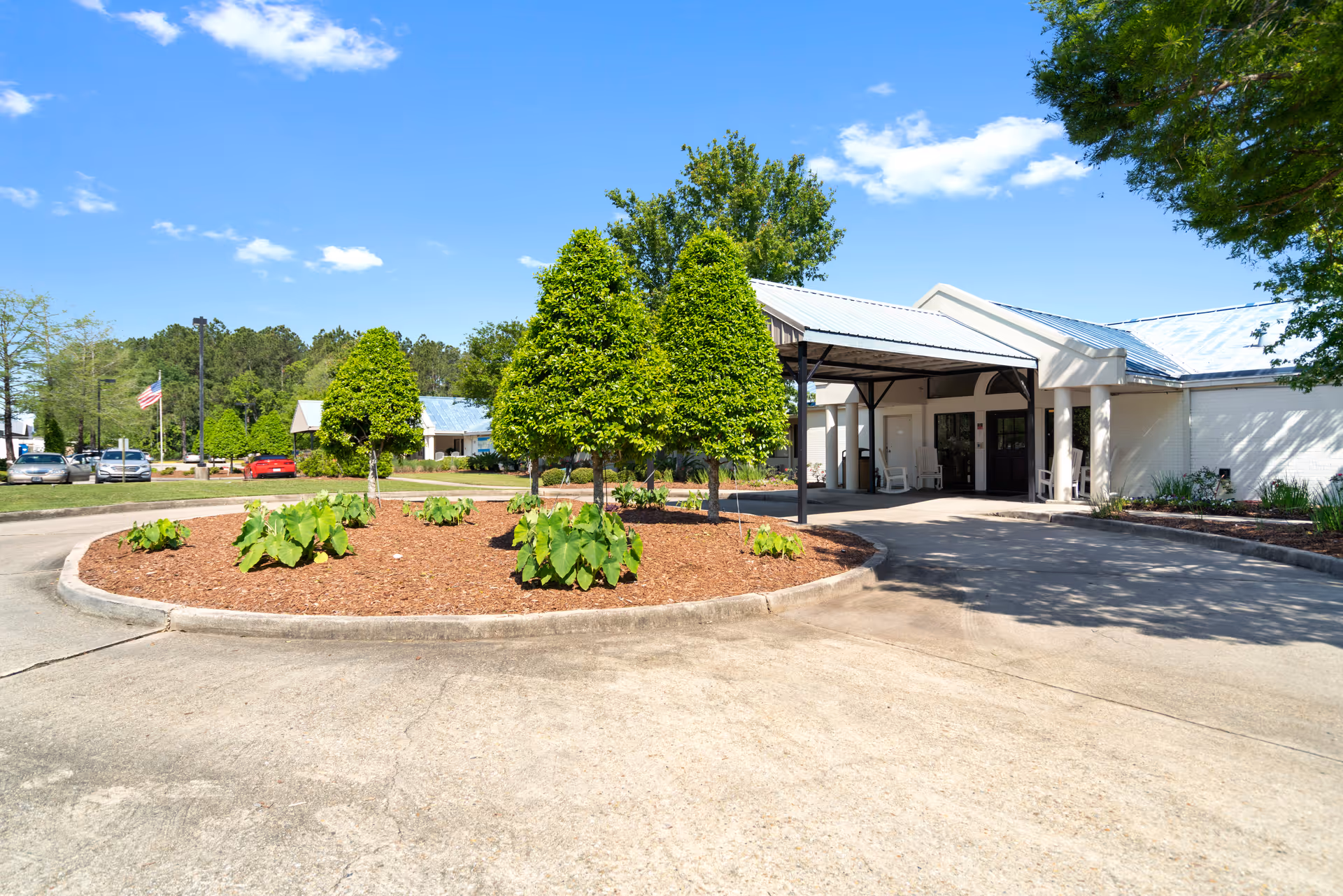 Front entrance of a single-story facility with a covered drop-off canopy, circular landscaped island, and parked cars under a clear blue sky.