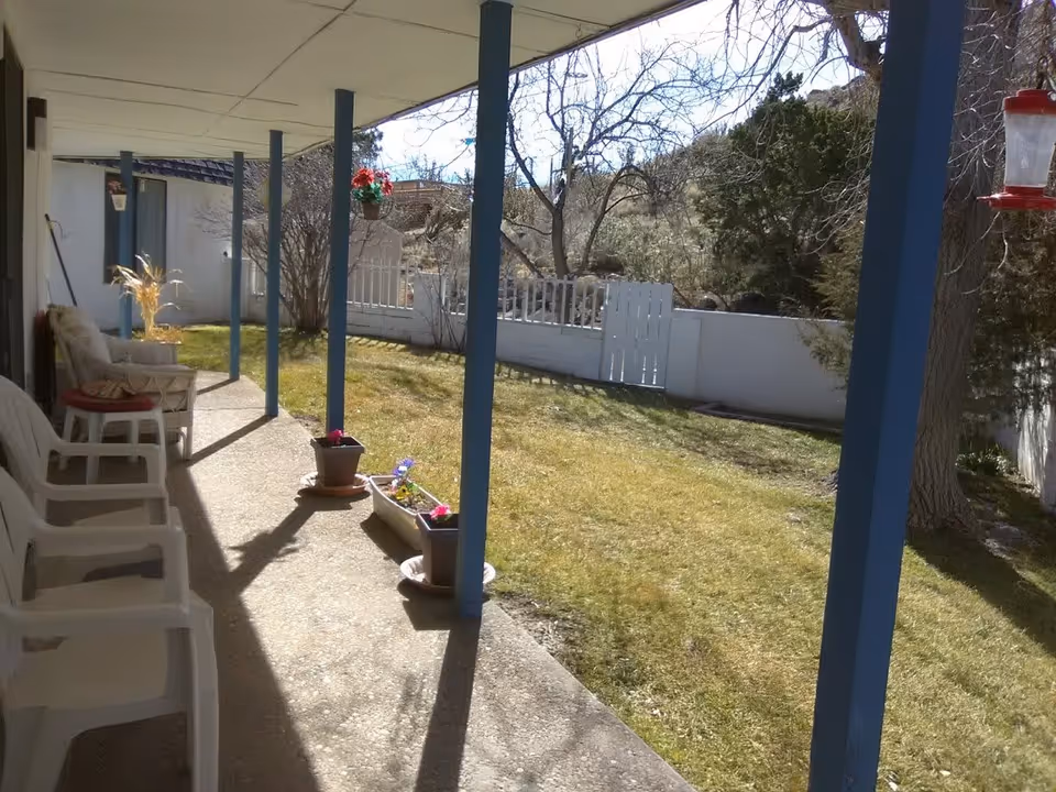 Covered patio area with white plastic chairs and potted flowers, overlooking a grassy yard with trees and a white fence under a clear sky.