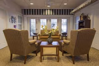 A cozy living room area with two patterned armchairs facing a wooden coffee table with a vase of flowers. Behind the table is a sofa with cushions, and large glass doors with white frames and striped valance letting in natural light.