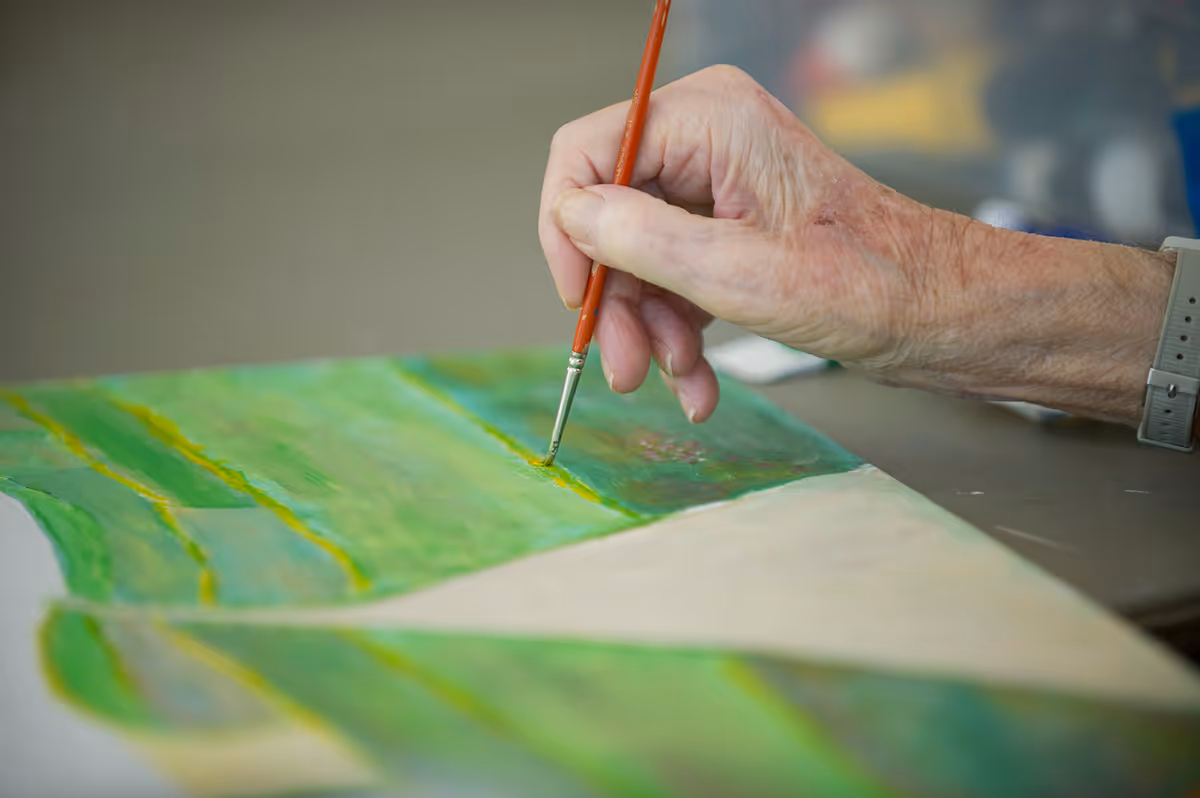 Close-up of an elderly person's hand holding a paintbrush and painting on a canvas with green and yellow colors.