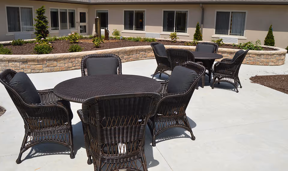 Outdoor patio area with two round tables surrounded by black wicker chairs with cushions, set on a concrete surface. Behind the patio is a low stone wall with planted shrubs and small trees, and the exterior wall of a building with several windows.