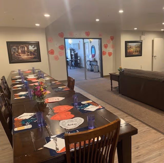 A long dining table set with placemats, napkins, utensils, and purple drinking glasses in a senior living facility. The table has heart-shaped paper decorations and a small flower arrangement. The room opens into another area with additional seating and wall decorations, including framed paintings and heart-shaped notes on the wall.