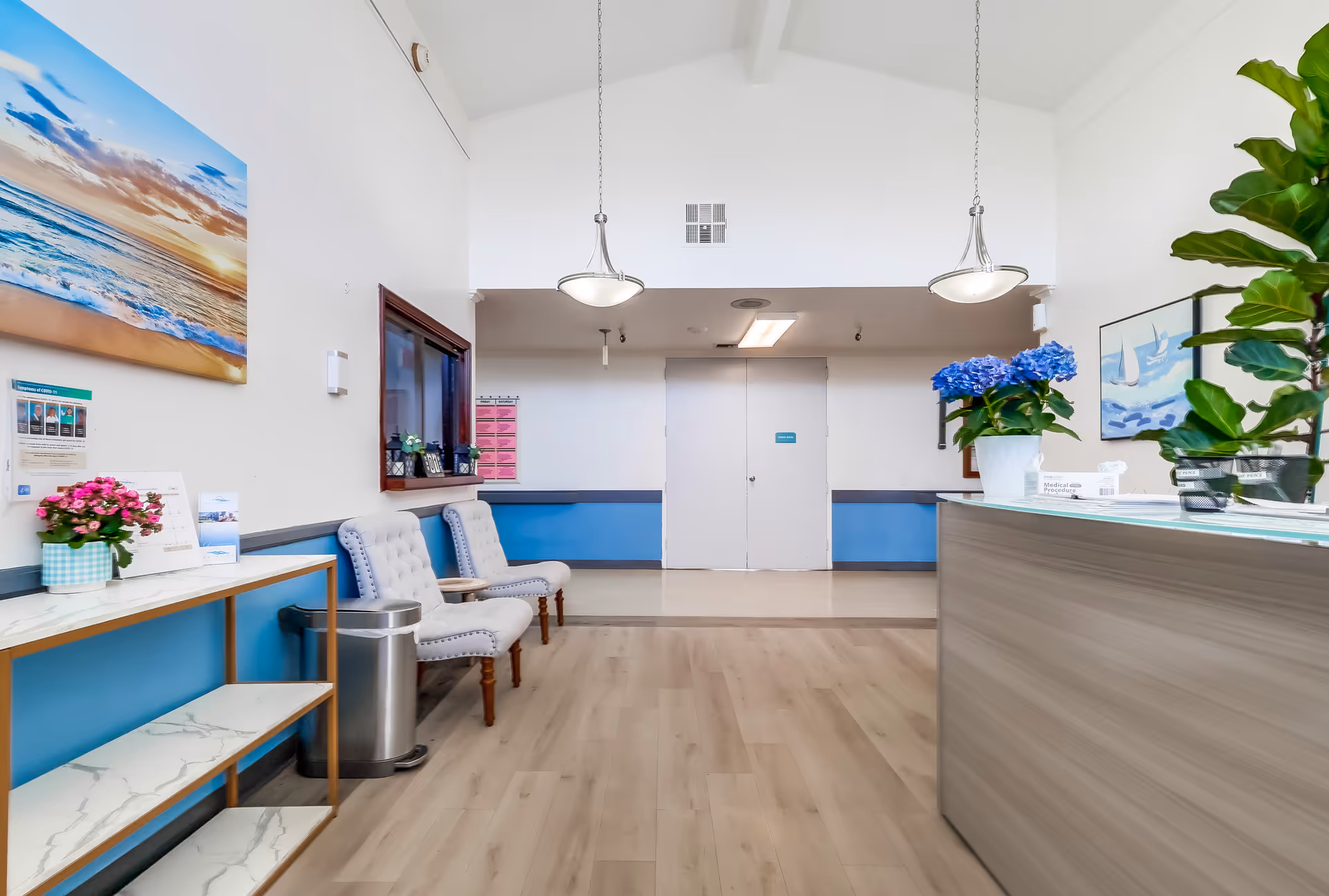 A bright and clean reception area with light wood flooring, two hanging pendant lights, and white walls with blue accents. There are two upholstered chairs along the left wall next to a small table with a pink flower pot and informational brochures. On the right side, there is a reception desk with a blue hydrangea plant and other greenery. The back wall has double doors and a small window with a counter beneath it. Artwork depicting a beach scene and sailboats decorates the walls.