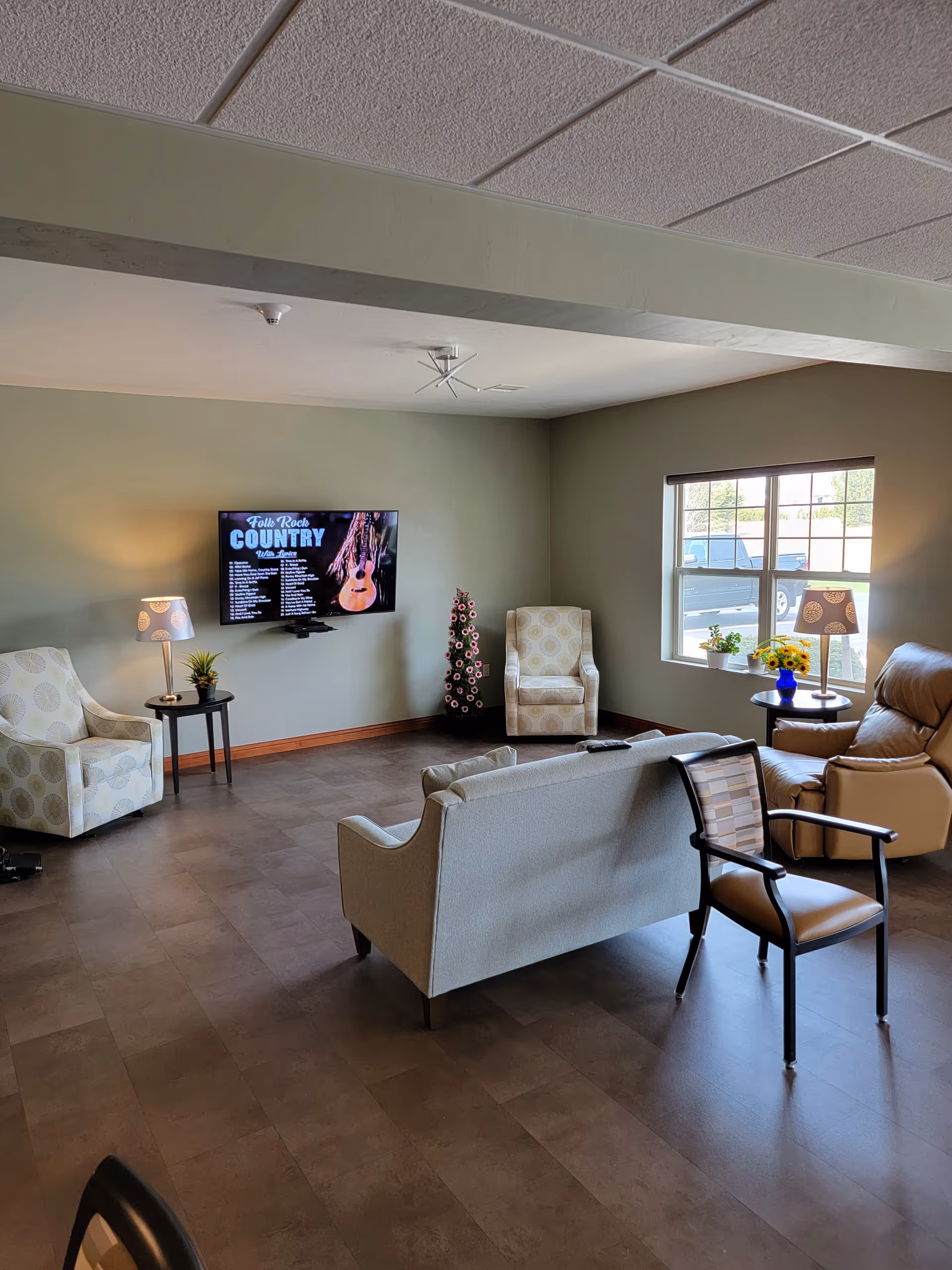 A cozy senior living facility common area with a wall-mounted TV displaying a country music playlist. The room has a beige loveseat, two patterned armchairs, a leather recliner, and a wooden chair arranged around the TV. There are two side tables with lamps and small plants, a window letting in natural light, and a decorative flower arrangement in the corner.