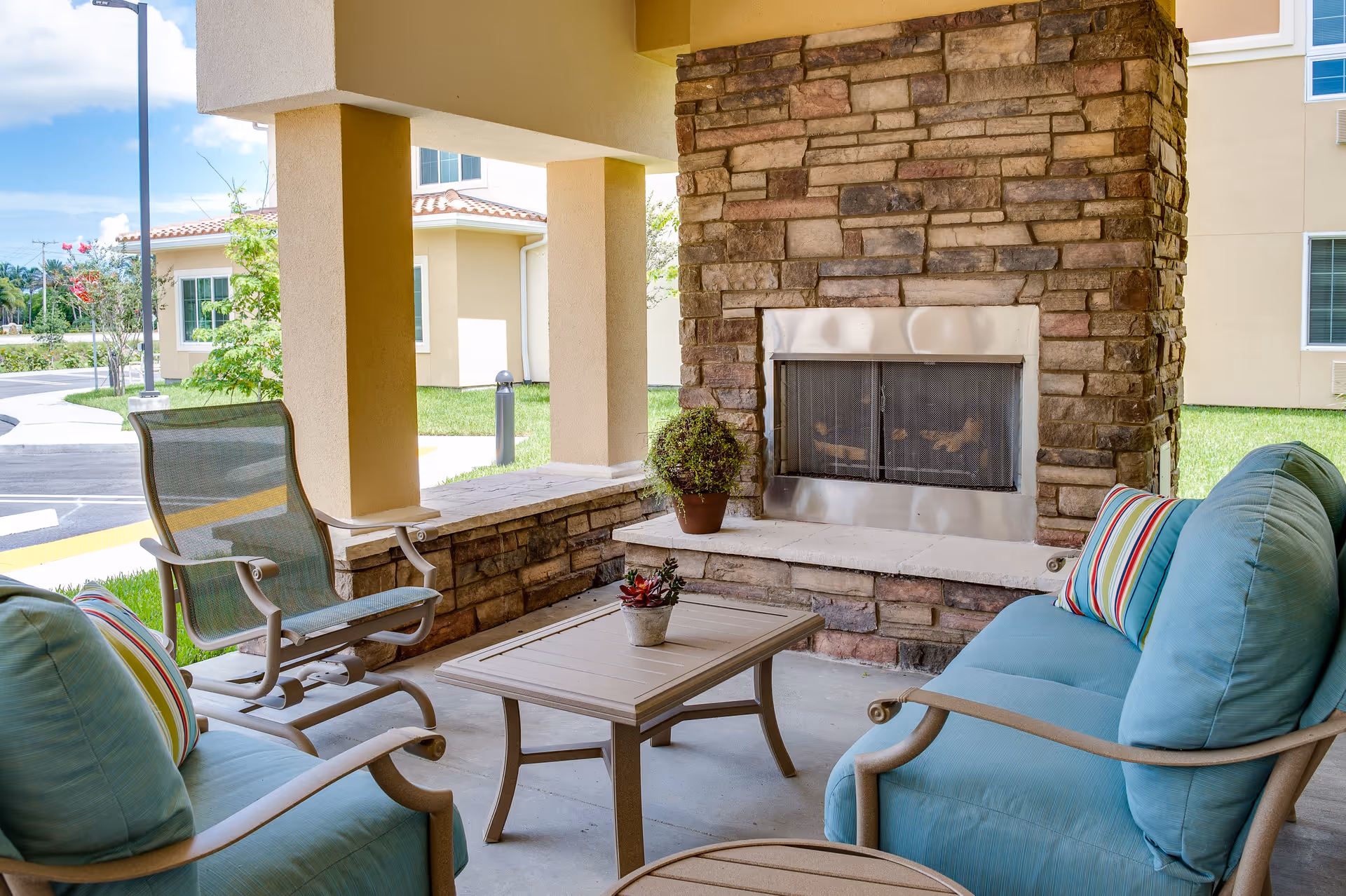 Outdoor covered seating area with a stone fireplace, two blue cushioned chairs with striped pillows, a mesh chair, and a small wooden table with potted plants. The area is adjacent to a building with beige walls and windows, and there is a parking lot and greenery visible in the background.