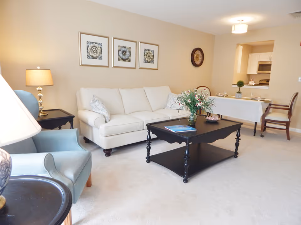 A cozy living room with a white sofa, two light blue armchairs, a black coffee table with a vase of flowers and a book, a side table with a lamp, and a dining area with a table set for two. The walls are beige with three framed floral artworks and a round wall clock. A kitchen area is visible through a pass-through window.