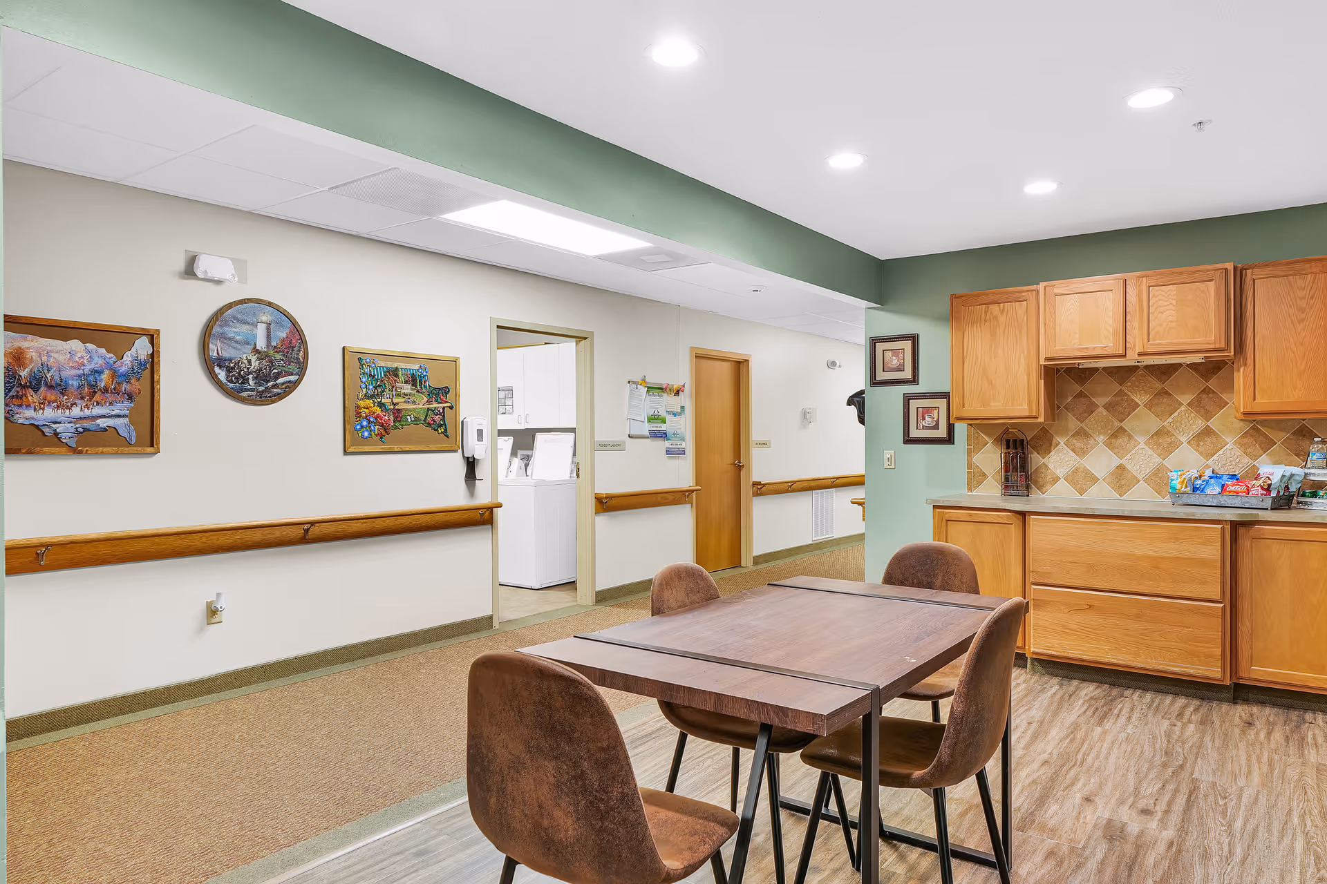 Interior view of a common area in an assisted living facility featuring a wooden table with four brown chairs, wooden cabinets with a tiled backsplash, and a hallway with doors and framed artwork on the walls.