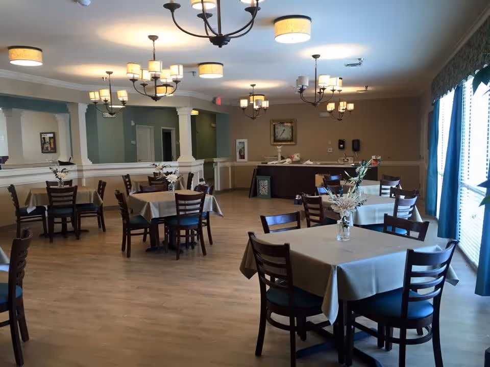 Bright dining room with square tables draped in beige tablecloths, wooden chairs, floral centerpieces, chandeliers, and large windows with curtains.