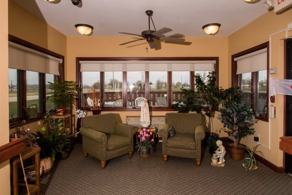 A cozy sitting area inside Prairie View Manor with two green armchairs facing each other, a small table between them holding a statue and a pot of pink flowers. The room has large windows with white blinds, several potted plants, beige walls, and ceiling lights with a ceiling fan.