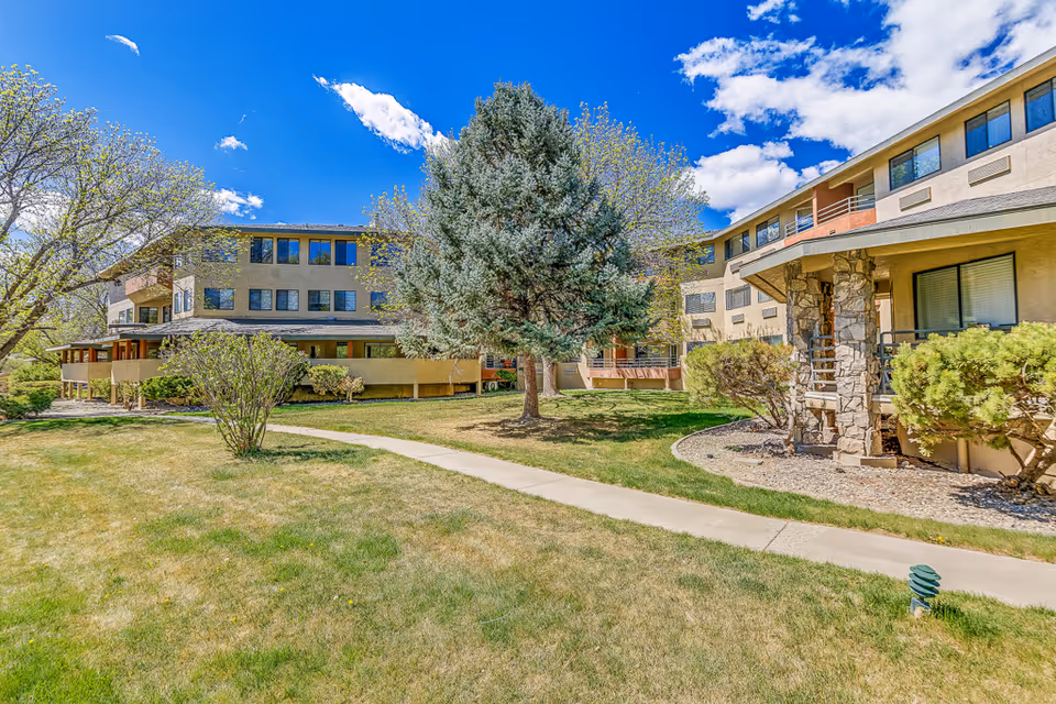 Exterior view of a senior living facility with a well-maintained lawn, a large tree in the center, and a paved walkway leading through the grassy area. The building has multiple windows and a stone pillar entrance under a clear blue sky with some clouds.