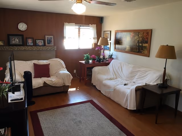 A cozy living room with two couches covered in white throws, a wooden side table with a lamp, a wooden console table with plants and a lamp, a window with white curtains, a ceiling fan with a light, framed pictures on the walls, and a rug on the wooden floor.