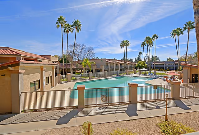 Outdoor swimming pool area surrounded by a fence with lounge chairs and umbrellas, tall palm trees, and senior living facility buildings in the background under a blue sky with some clouds.