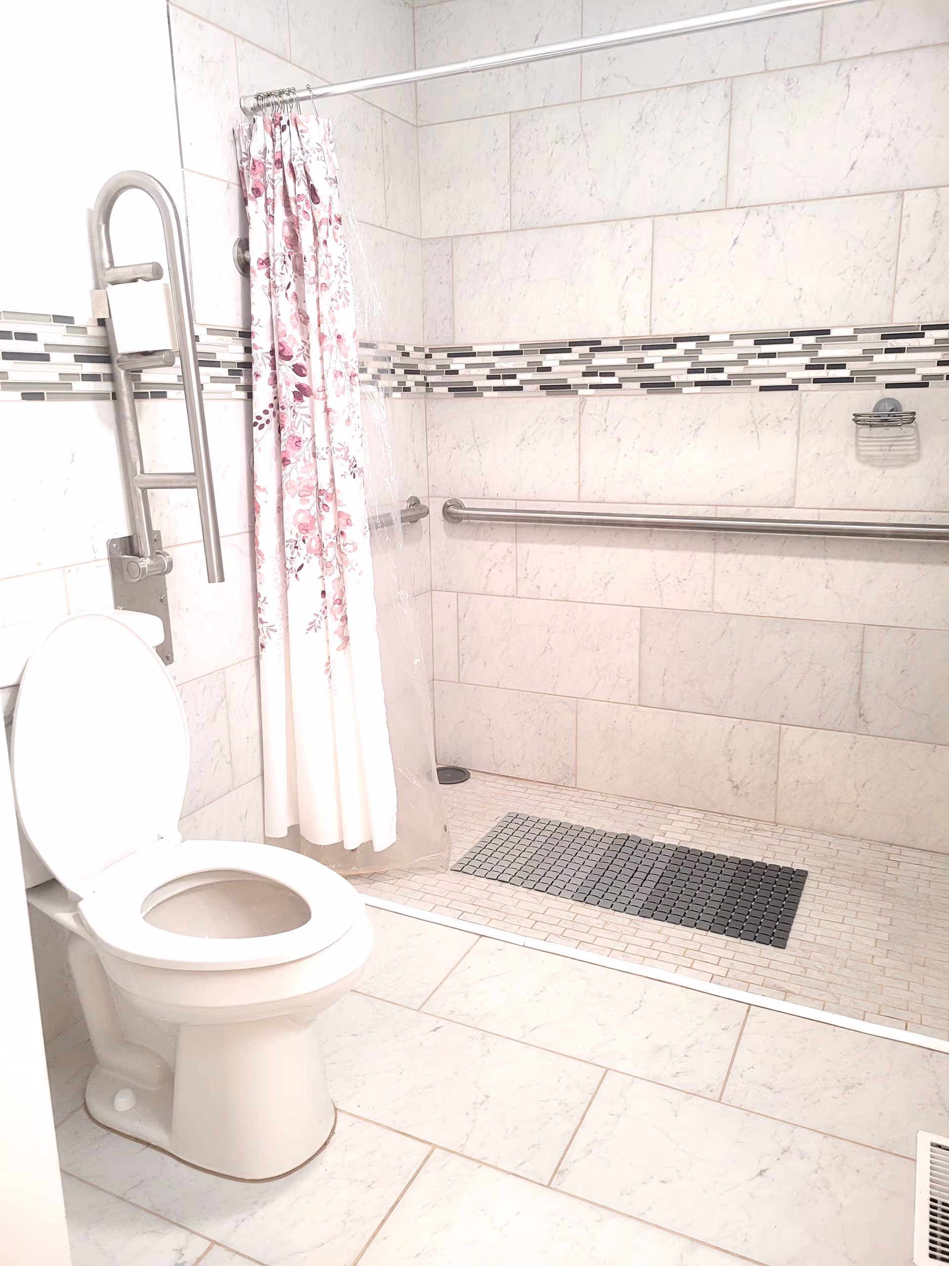 A clean bathroom with white tiled walls and floor. There is a white toilet with the lid open on the left side. Next to the toilet is a metal grab bar for support. The shower area has a floral-patterned shower curtain and a clear plastic liner. The shower floor has small square tiles with a darker rectangular mat in the center. A horizontal grab bar is mounted on the shower wall for safety.