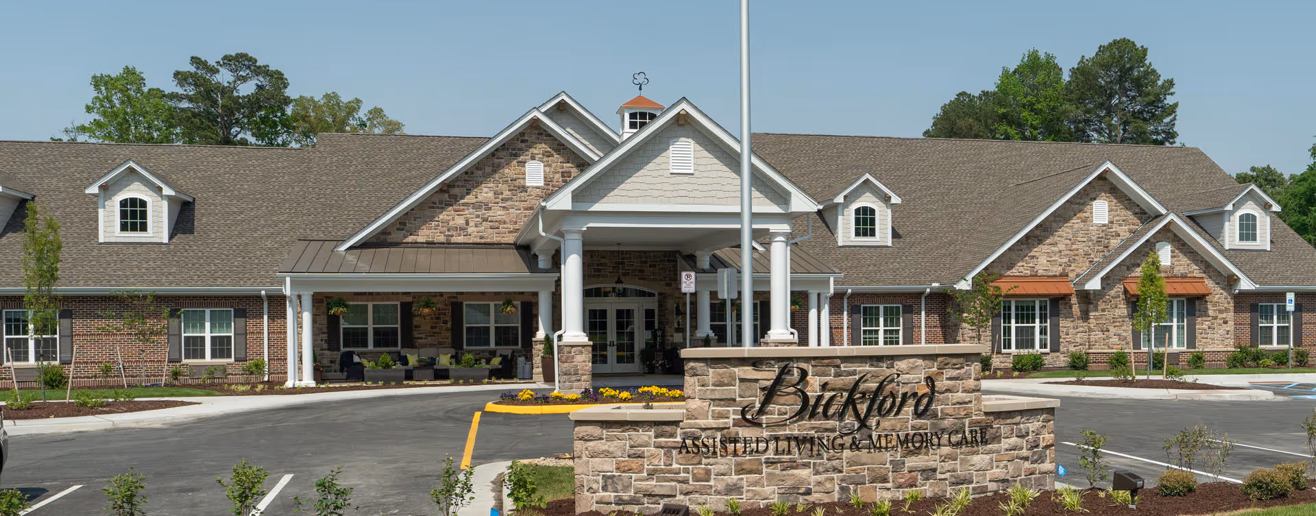 Front exterior view of Bickford of Chesapeake, an assisted living and memory care facility with a large entrance, stone and brick facade, and a landscaped parking area.
