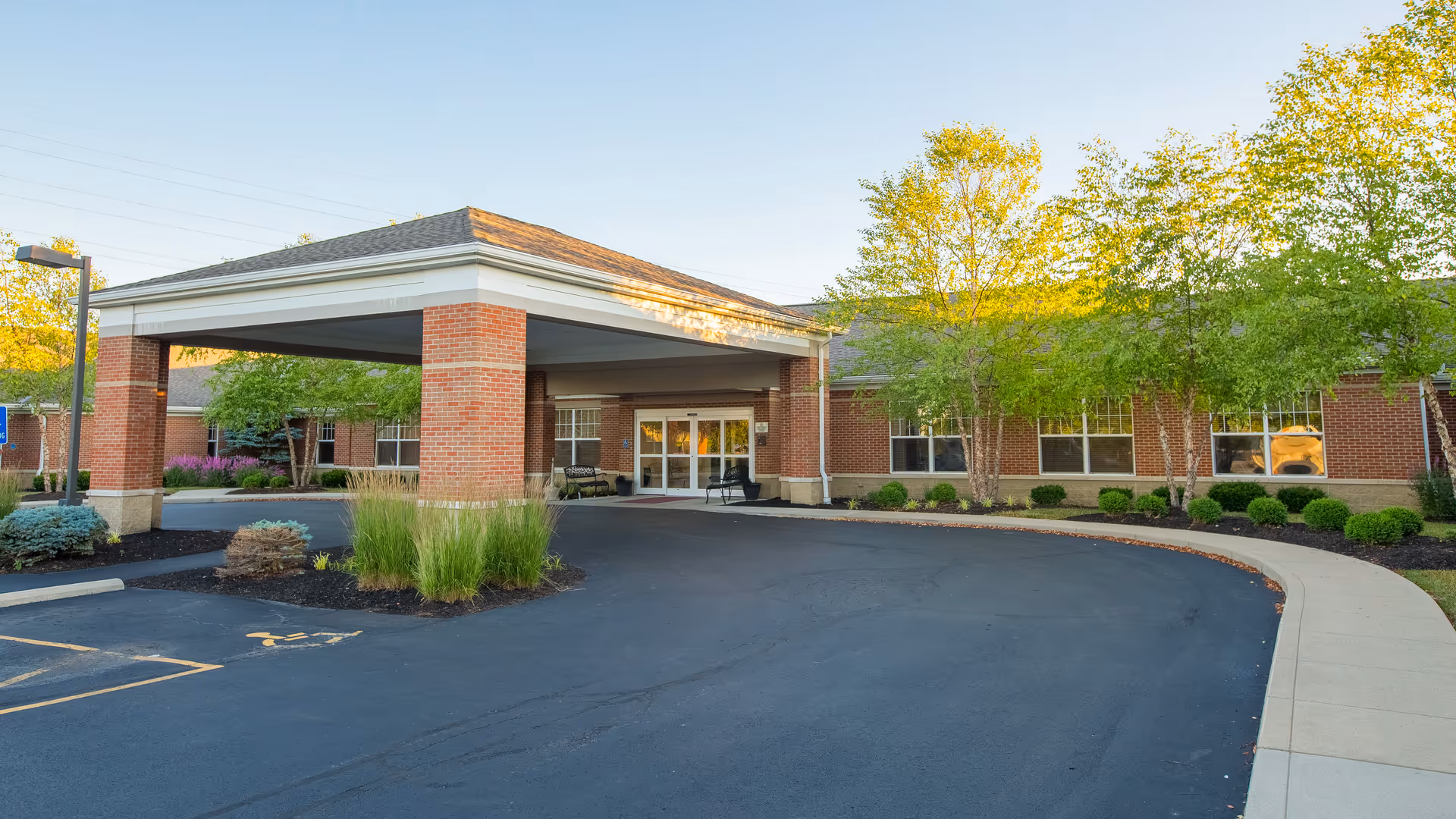 Exterior view of Tuscany Gardens senior living facility entrance with a covered drop-off area, brick pillars, surrounding greenery, and a paved driveway.