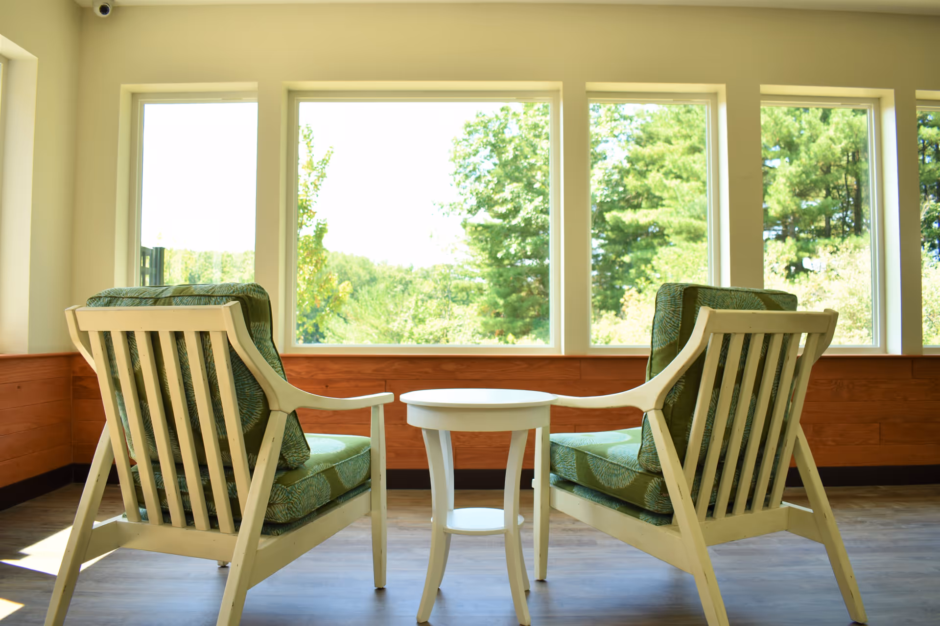 Two wooden armchairs with green cushions facing a small round white table in front of large windows showing a view of green trees outside in a bright room.
